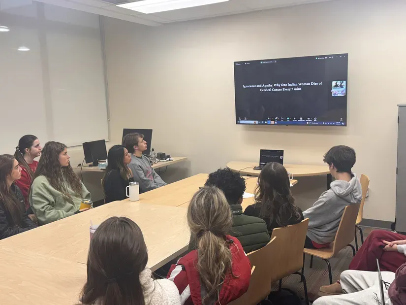 High school students sit around a long table and look at a screen, where a zoom meeting connects them to journalist Swagata Yadaver